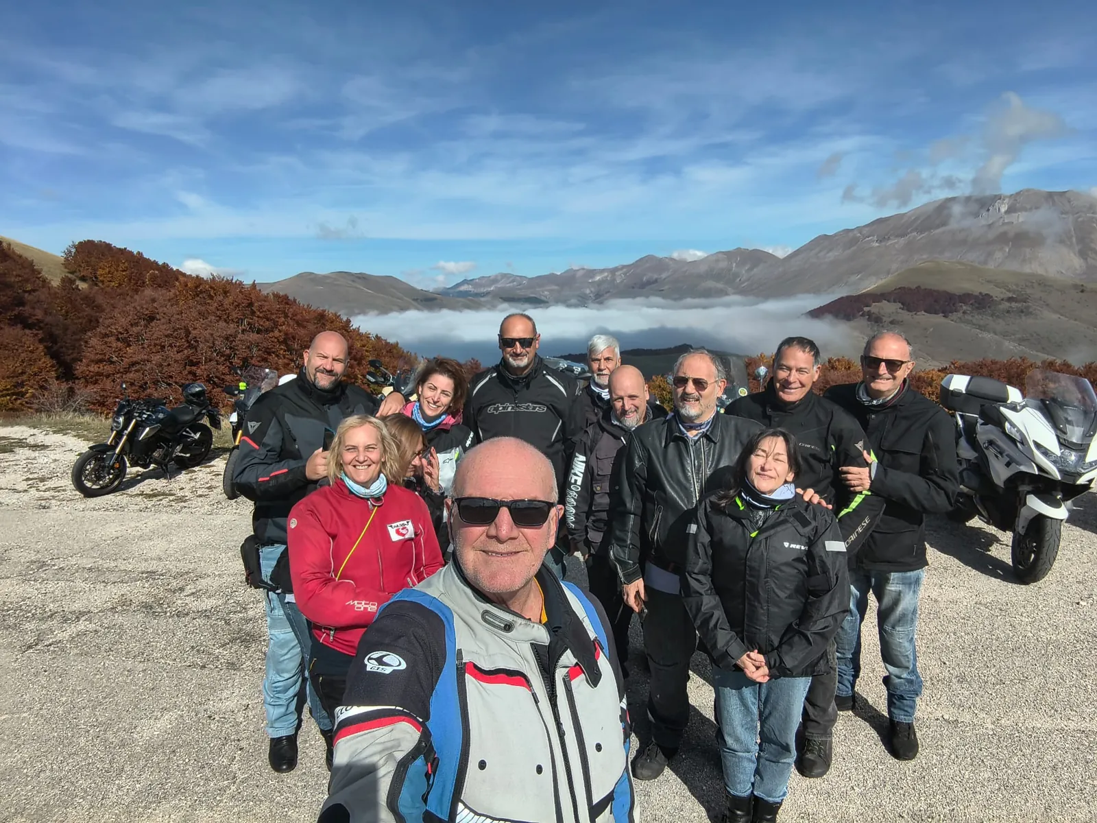 Selfie di gruppo di motociclisti sorridenti in montagna, con le moto parcheggiate e le nuvole in valle
