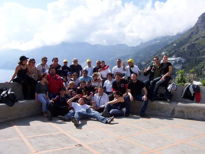 Foto di gruppo di una trentina di motociclisti seduti su un muretto sul mare, con le montagne alle spalle
