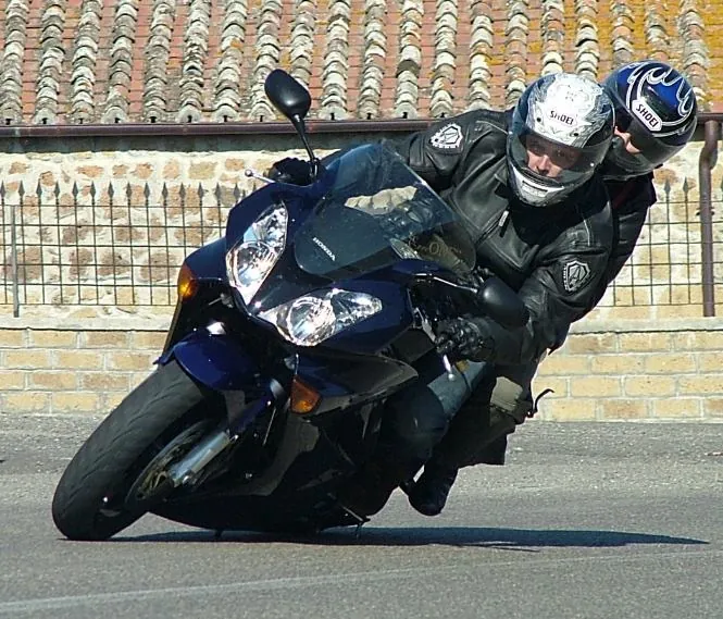 Una moto sportiva blu in piega con pilota e passeggero, davanti a un muro di mattoni